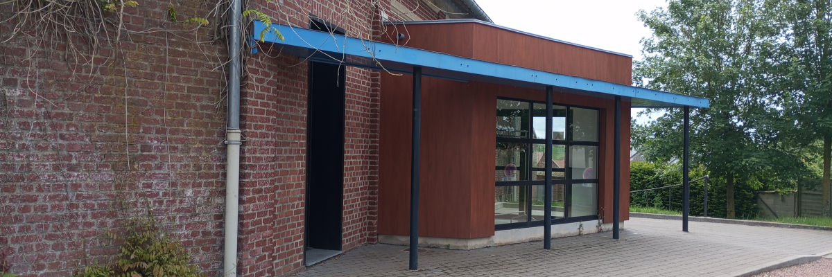 Bâtiment en brique avec entrée vitrée, auvent bleu, allée pavée et végétation.