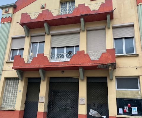 Foyer municipal: bâtiment jaune avec portes et fenêtres métalliques, affronté par une route mouillée.