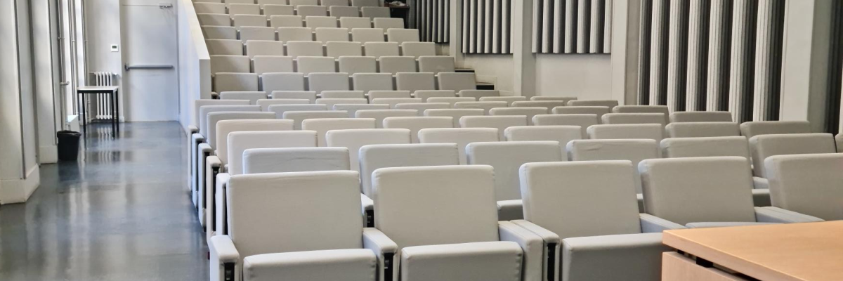 Salle de classe avec rangées de chaises, table de professeur, tableau blanc et éclairage suspendu.