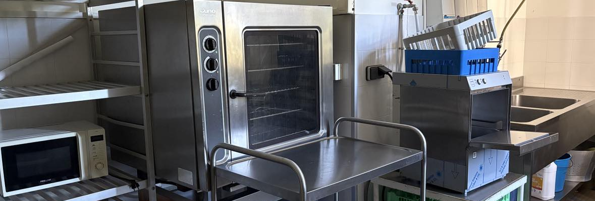 Cuisine avec four industriel, micro-ondes, étagères, chariots, lave-vaisselle et plateaux.