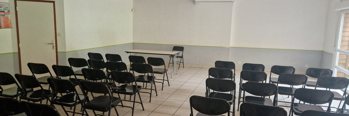 Salle avec rangées de chaises, table, porte, chariot rouge et blanc en hauteur, éclairage au plafond.