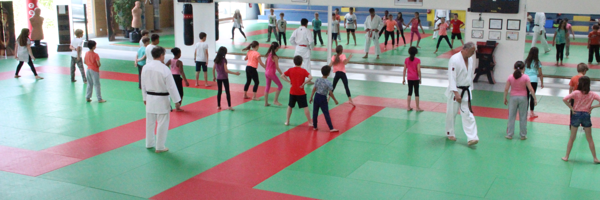Salle de sport avec tatamis, groupes d'enfants et adultes en tenue de sport, écrans d'affichage et horloges.