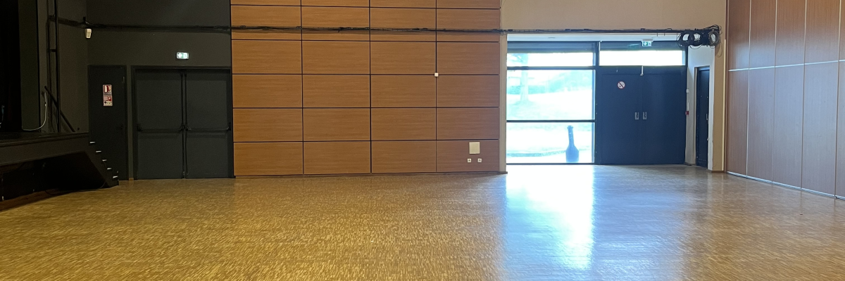 Salle spacieuse avec plancher en bois, murs en bois, éclairage au plafond, rideau de scène, portes vitrées et systèmes audio/son.