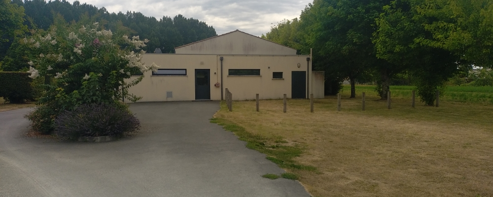 Bâtiment avec entrée, porte, fenêtres et parking. Arbres et buissons autour.