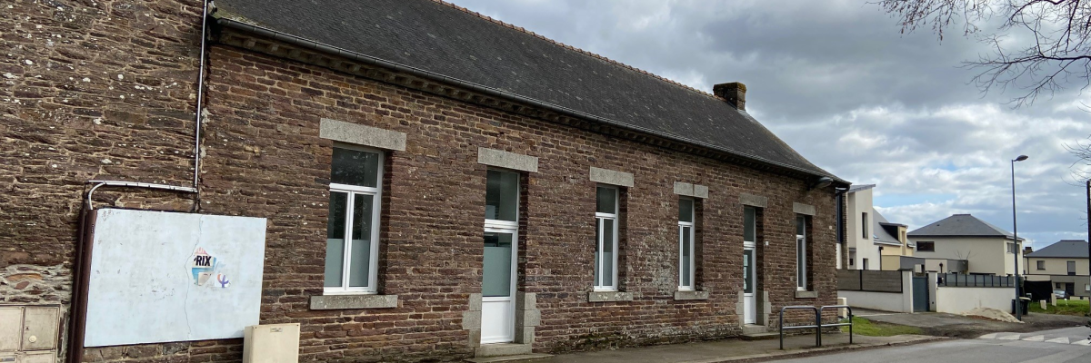 Un bâtiment en brique à un étage avec des fenêtres blanches, un panneau, une route avec un passage piéton, des arbres sans feuilles.