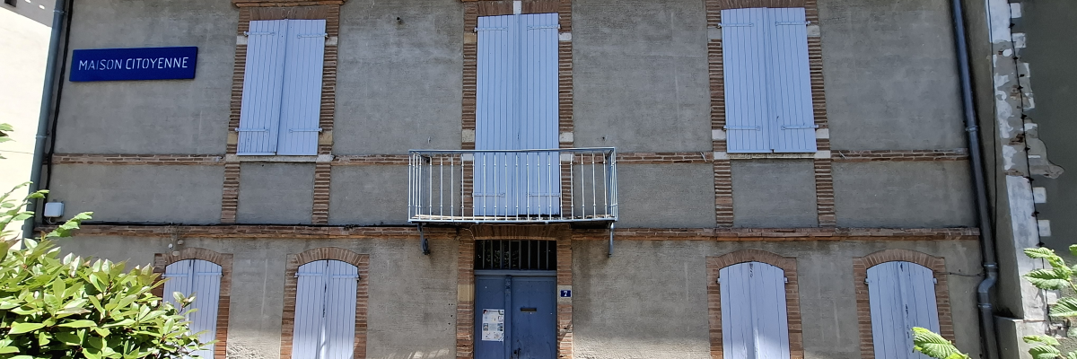 Bâtiment avec façade en briques, volets gris, balcon métallique, porte bleue et panneau "Maison Citoyenne".