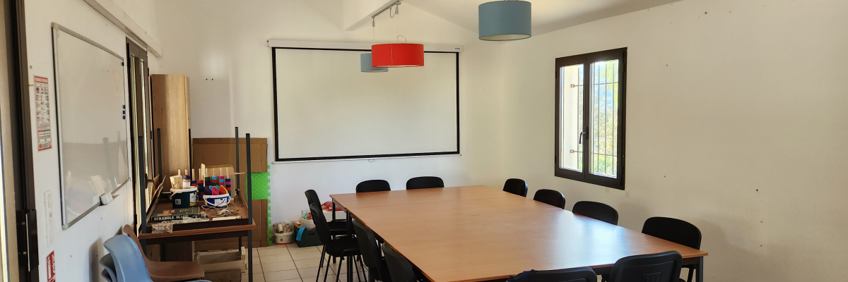 Salle avec table, chaises, tableau blanc, écran, ventilateur, et étagères.