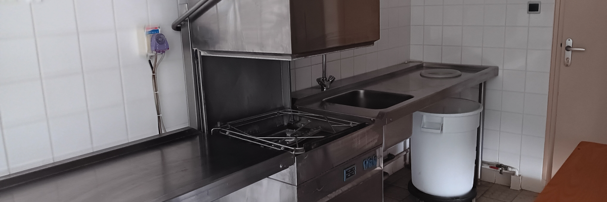 Cuisine avec lave-vaisselle industriel, plans de travail en inox, évier et bidon.