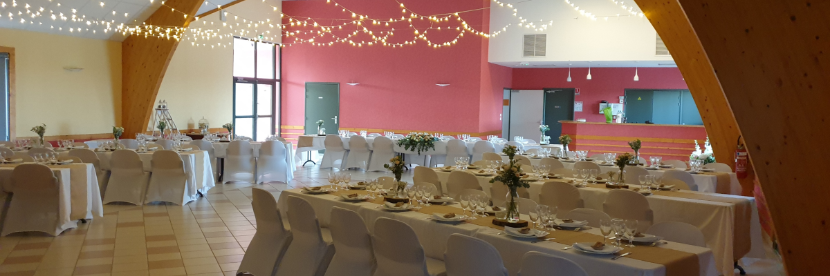 Salle avec tables, chaises, nappes, guirlandes lumineuses, vaisselle et fleurs.