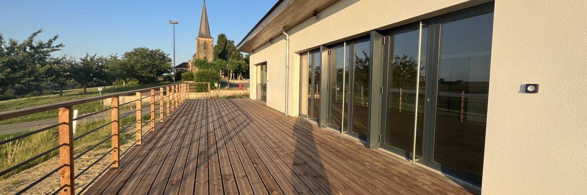 Terrasse en bois menant à un bâtiment avec fenêtres vitrées. Eglise lointaine et ciel dégagé.