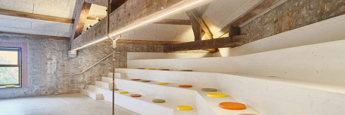 Salle avec gradins, rampes, poufs et poutres apparentes.