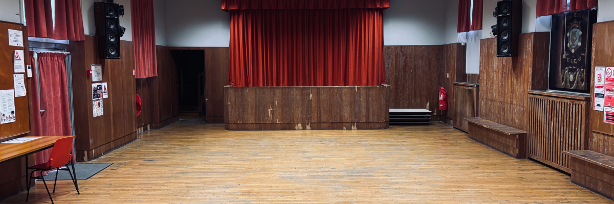 Salle avec scène, rideaux rouges, haut-parleurs, bancs et porte.