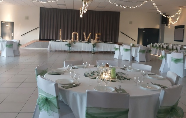 Salle de réception avec tables, chaises, nappes, vaisselle, guirlandes lumineuses et décorations florales.