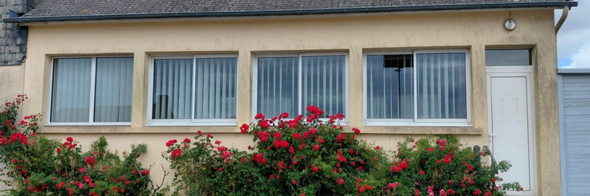 Maison beige avec toit gris, trois fenêtres, porte blanche, et buissons de fleurs rouges.