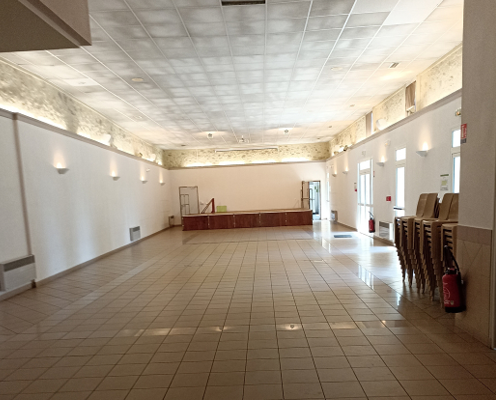 Salle vide avec sol carrelé, murs blancs, luminaires, et chaises empilées.