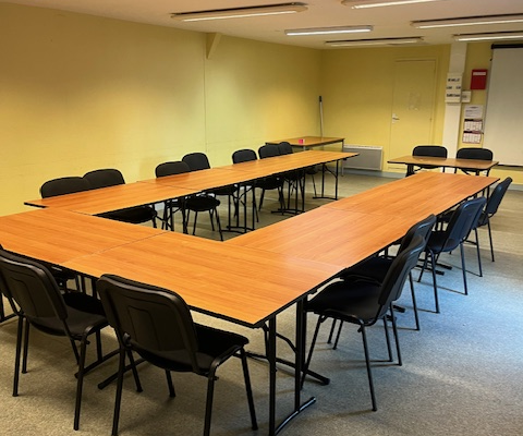 Salle avec tables disposées en U, chaises, éclairage et armoire métallique.