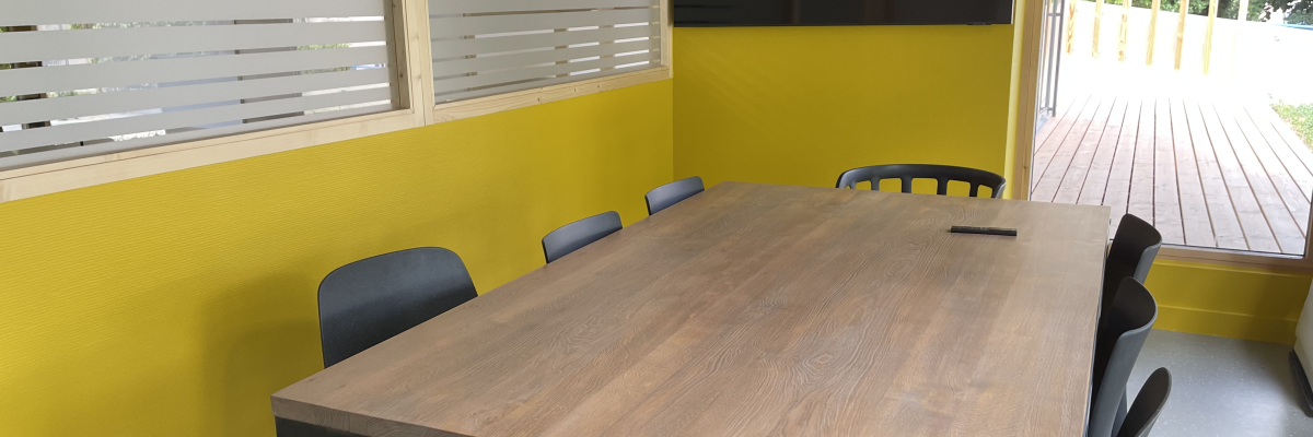 Table de réunion avec six chaises, mur jaune, fenêtres avec stores et porte ouverte.
