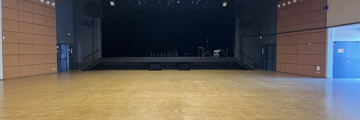 Salle vaste avec plancher en bois, scène, éclairages, murs en bois et portes bleues.