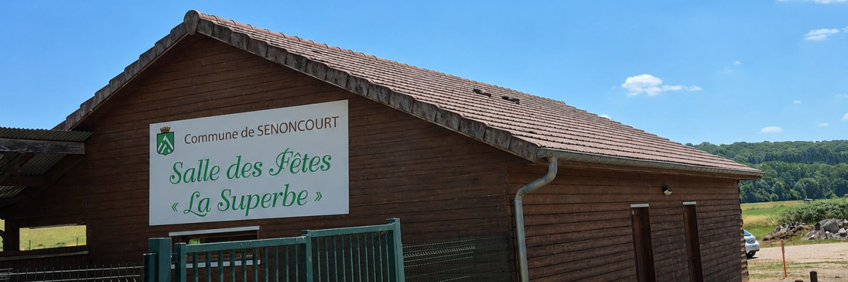 Bâtiment en bois avec une pancarte indiquant "Salle des Fêtes La Superbe" et un portail vert.