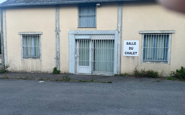 Bâtiment avec trois fenêtres grillagées et une porte centrale. Ensoleillé, devant une route.