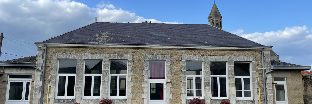 École en pierre avec fenêtres, portes, toit, et un clocher. Cour avec banc et fleurs.