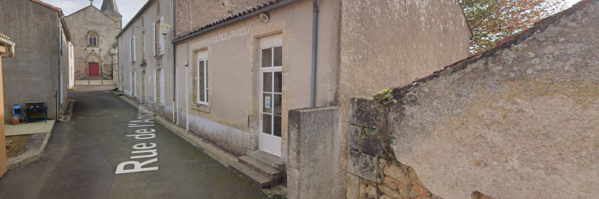 Rue étroite avec bâtiments anciens en pierre, église au fond, poteaux électriques, arbres dénudés.