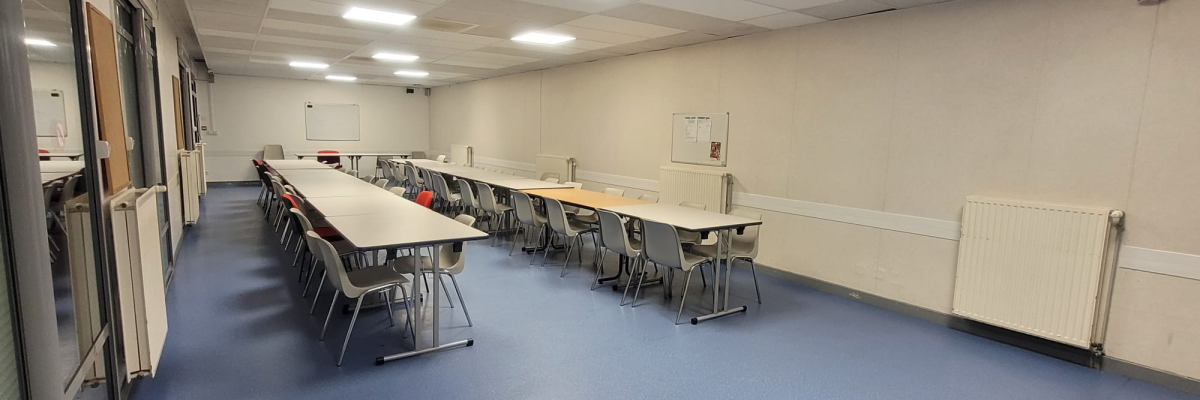 Salle de classe avec tables, chaises, tableau blanc et radiateurs.