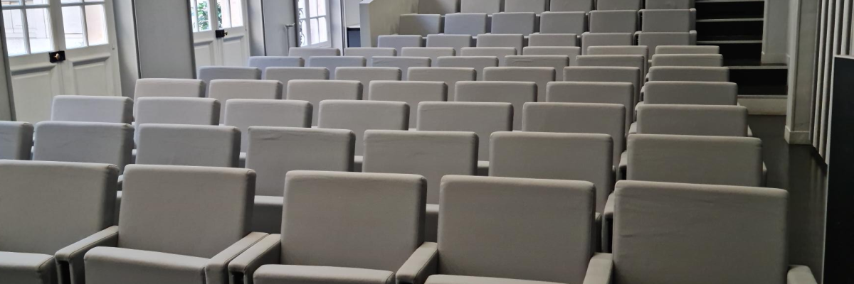 Salle de cours avec rangées de chaises, table devant, ordinateur, clavier, souris et documents.