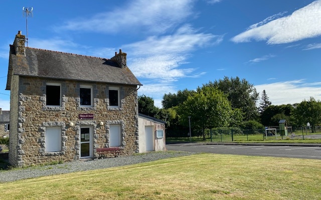 Maison en pierre avec toit en ardoise, antenne sur le toit, banc rouge devant, pelouse et route.