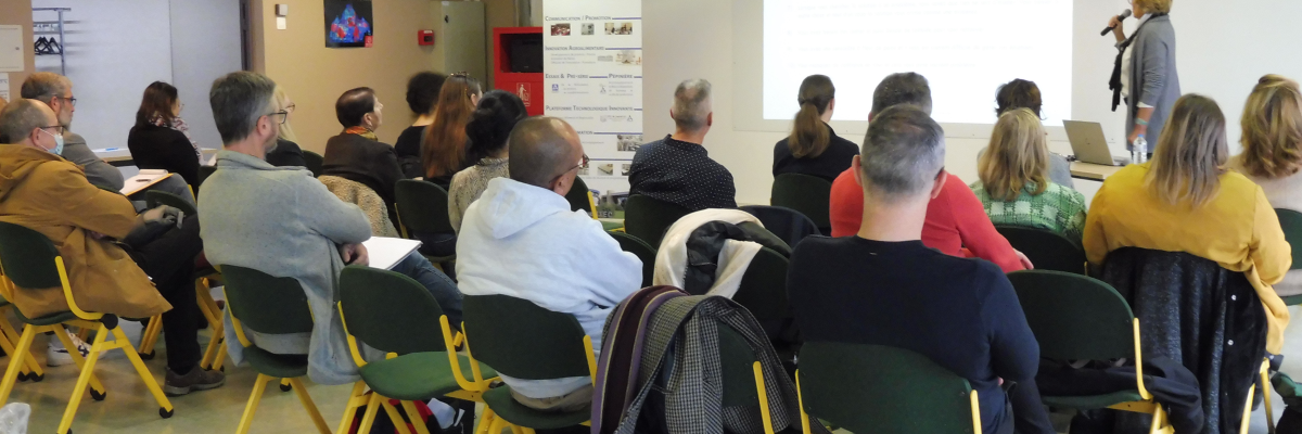 Salle de réunion avec projecteur, écran, table, chaises, un présentateur et un public.