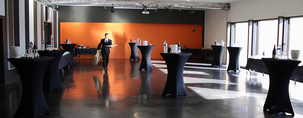 Salle avec tables hautes, projecteur, écran orange et fenêtres.