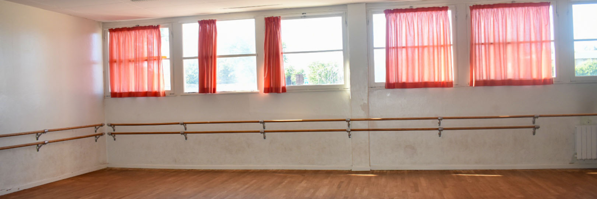 Salle de danse avec parquet, barres murales, fenêtres et rideaux rouges.