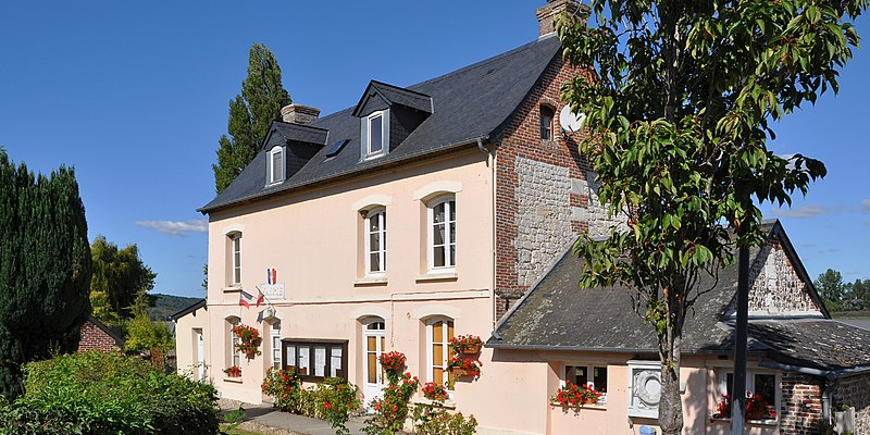 Maison à deux étages avec toit noir, murs beiges et pierres apparentes. Fenêtres avec volets blancs. Jardinières avec fleurs rouges. Arbre à droite. Ciel bleu.