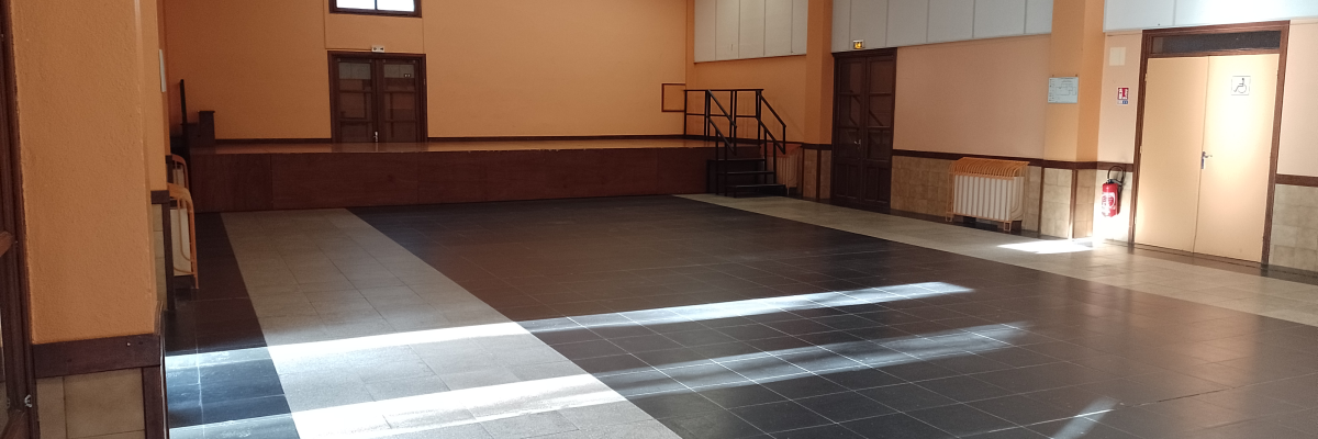 Salle spacieuse avec sol en carreaux de pierre, murs peints, plafond à poutres apparentes, fenêtres, portes, radiateurs, escaliers et extincteur.