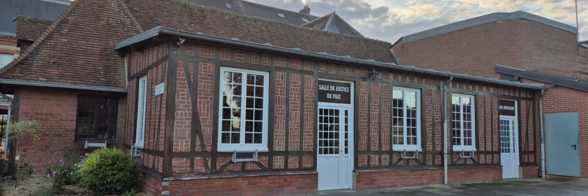 Bâtiment en briques avec toit en tuiles, fenêtres et portes blanches, "Salle de justice de paix" inscrite sur la porte. Cour asphaltée.