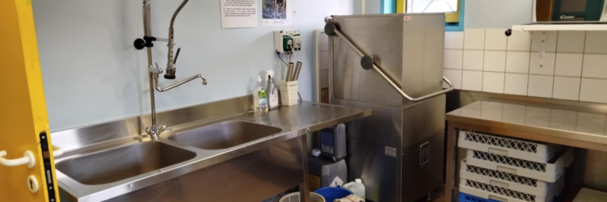 Cuisine industrielle avec évier, lave-vaisselle, chariot et produits de nettoyage.