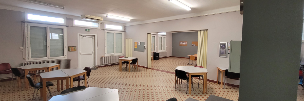 Salle de classe avec tables, chaises, tableau blanc, vidéoprojecteur, distributeur d'eau et radiateurs.