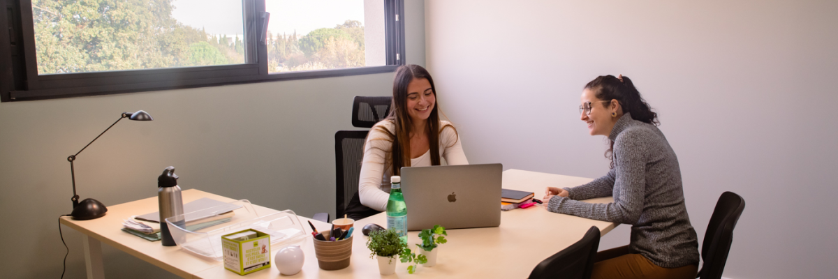 Bureau avec deux femmes, table, chaises, ordinateur portable, lampe de bureau, bouteille d'eau, plantes.