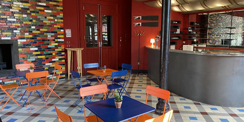 Salle avec tables, chaises, comptoir, et étagères. Mur coloré et sol carrelé.