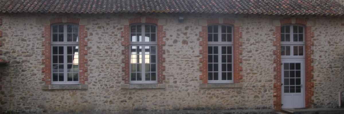 Bâtiment en pierre avec toiture en tuiles, quatre fenêtres et une porte blanche. Cour asphalte.
