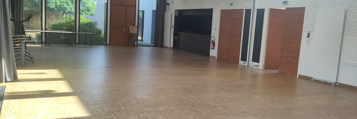Salle spacieuse avec parquet, portes en bois, fenêtres, luminaires suspendus, et mobilier de jardin visible à travers les portes vitrées.
