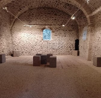 Pièce en pierre, sol en paillage, cubes en pierre, fenêtres vitrées avec motifs.