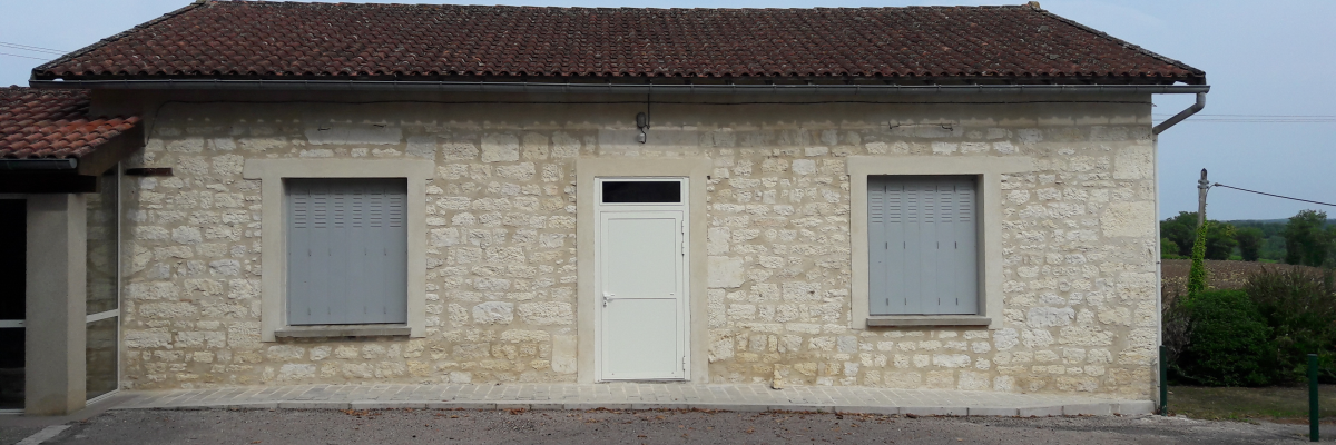 Maison en pierre, toit en tuiles, porte blanche, deux fenêtres, allée gravillonnée.