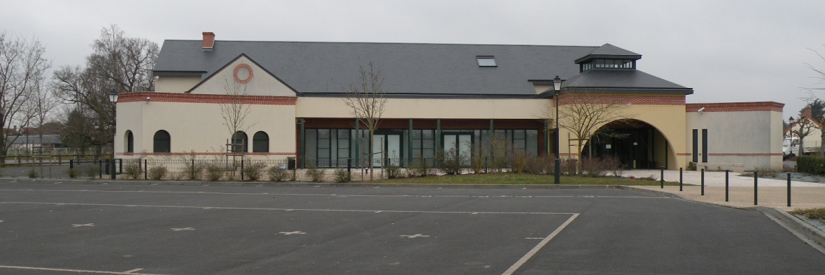 Bâtiment avec façade beige, toiture grise, fenêtres et porte d'entrée. Parking vide devant.