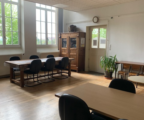 Salle avec tables, chaises, armoire, plantes vertes, horloge et fenêtres.