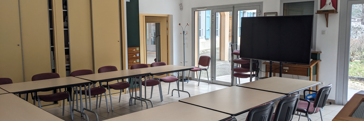 Salle de classe avec tables, chaises, tableau noir, écran, horloge, armoire.