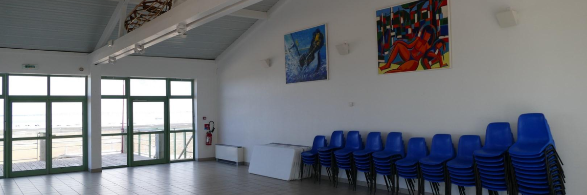 Salle avec chaises bleues, tableaux, porte et fenêtres sur un mur, sol carrelé.