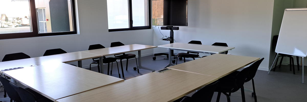 Salle de réunion avec table, chaises, tableau blanc, écran et fenêtres.