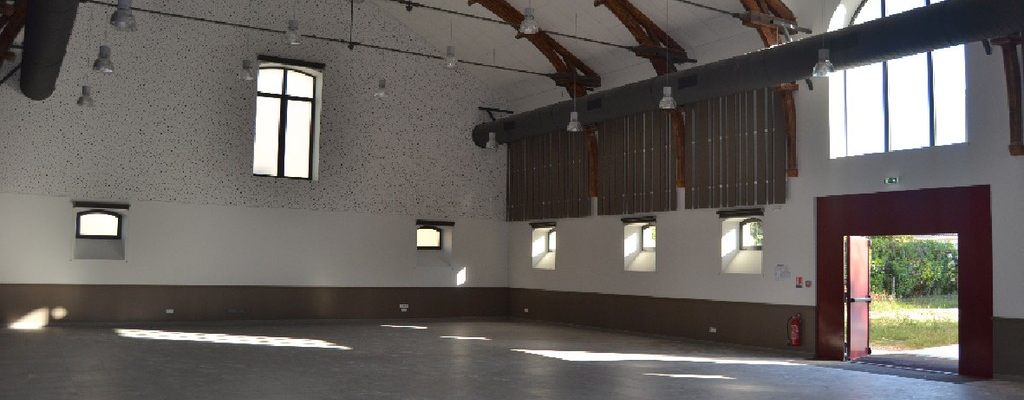 Grande salle avec plafond voûté en bois, murs blancs, fenêtres, porte, et sol en carreaux.