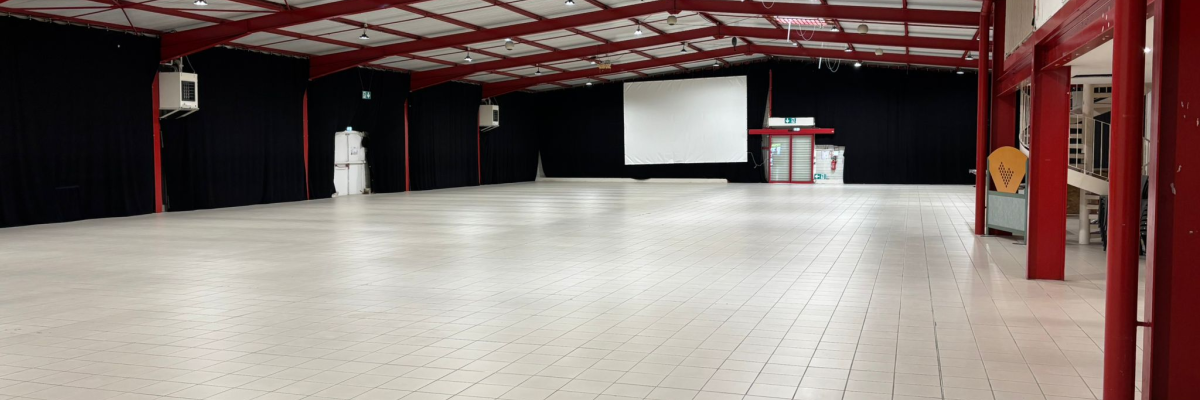 Salle spacieuse avec sol carrelé, piliers rouges, écran blanc, tableau d'affichage et distributeur automatique.